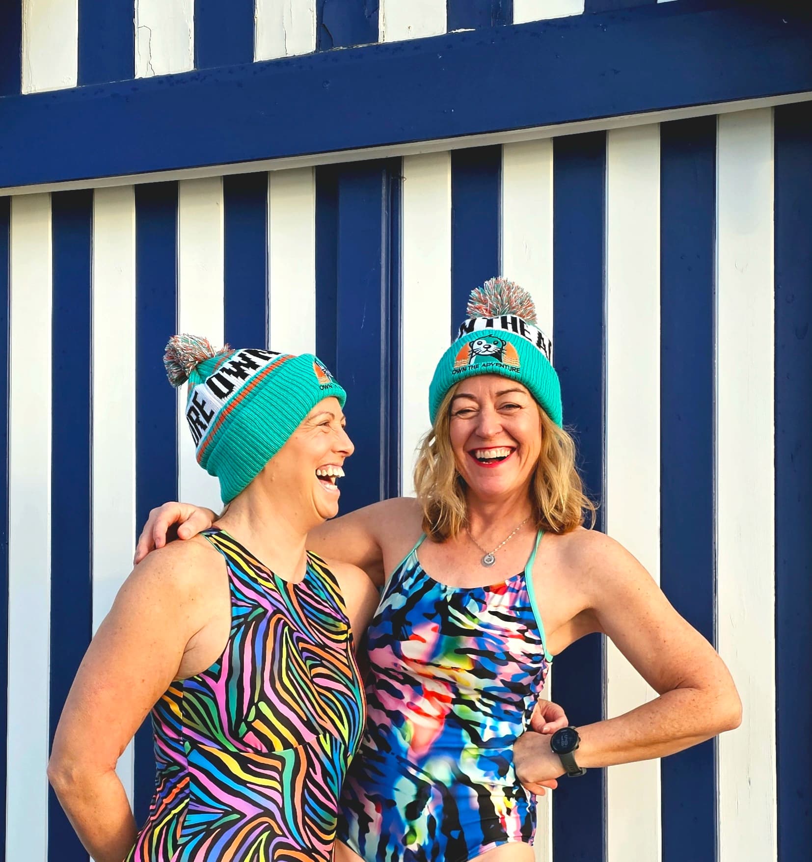 Two smiling swimmers in colourful swimwear beside a striped beach hut